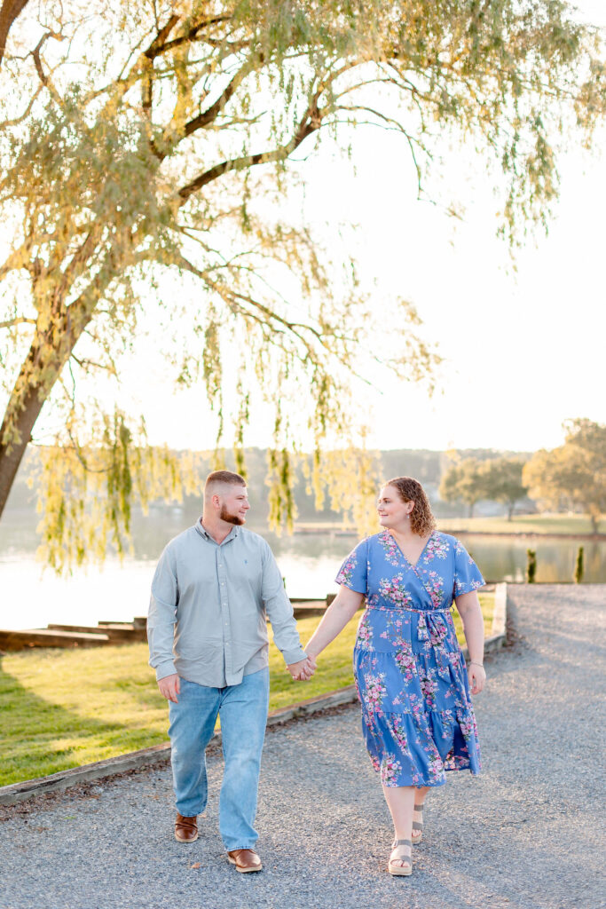 A Woman smiles while walking with her fiancee as they hold hands under a willow tree by the water